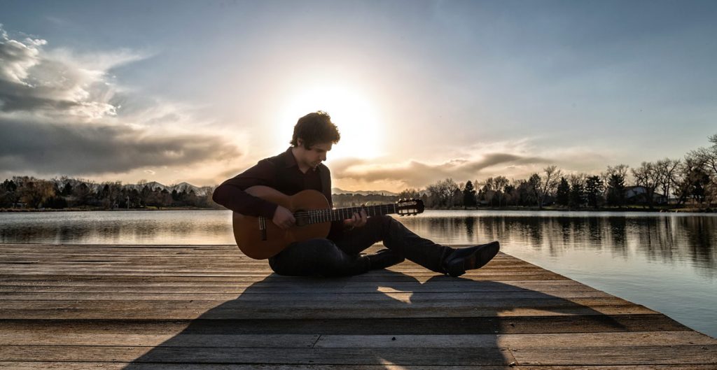 A classical guitarist sits on the dock of a lake at sunrise.