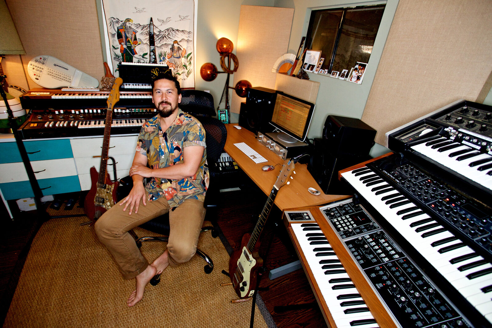 Mitchell Yoshida pictured in his home studio sitting on a chair surrounded by keyboards, guitars and recording equipment 