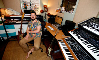 Mitchell Yoshida pictured in his home studio sitting on a chair surrounded by keyboards, guitars and recording equipment