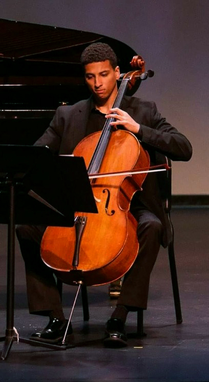 Myles Yeazell dressed in concert black playing cello on stage next to a piano