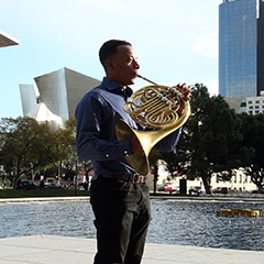 Malik Taylor plays the french horn in a plaza in downtown LA