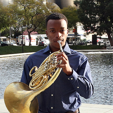 A musician plays the french horn and looks into the camera