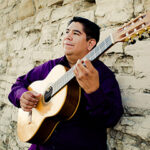 Tony Ybarra pictured holding a guitar beside a stone wall