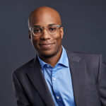 Jonathan Azu pictured in a suit and blue shirt, smiling to camera