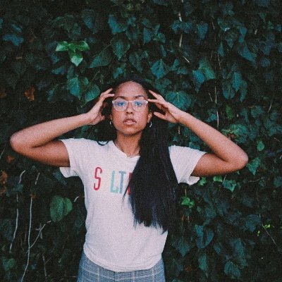 Jensen McRae holding hands to her face, standing in front of a greenery covered wall