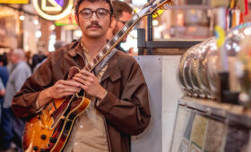 student holding an electric guitar with neon signs in the background.