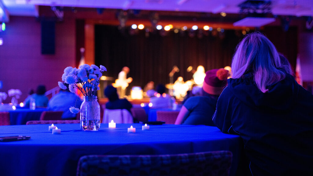 A person sits alone at a table with small candles plus a bouquet of flowers and watches a performance taking place at a distance onstage. 