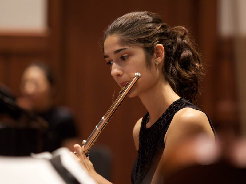This is a close-up view of a USC Thornton student in black concert attire playing the flute with the rest of the chamber orchestra. 