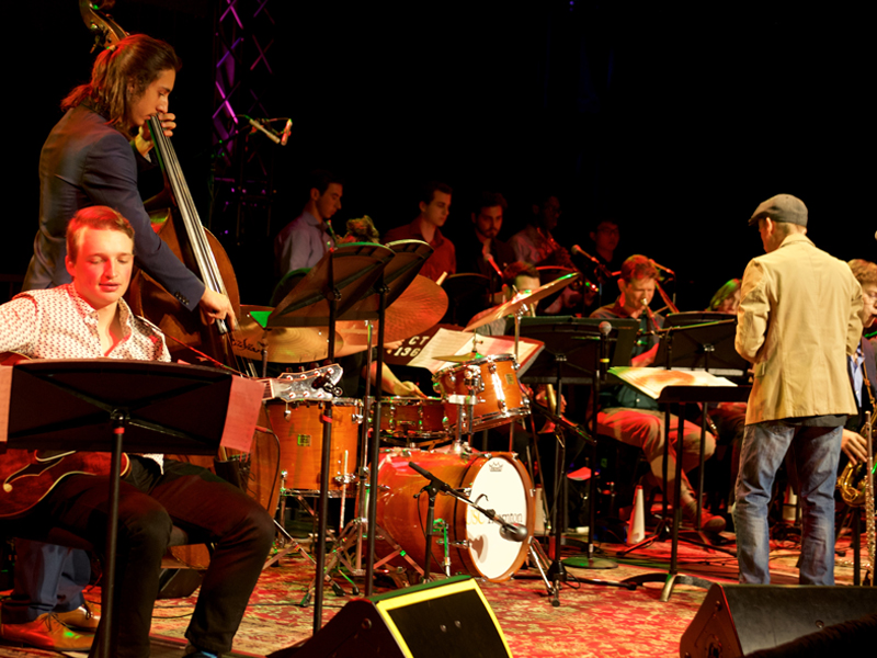 Members of the concert jazz orchestra in various attire perform onstage. 