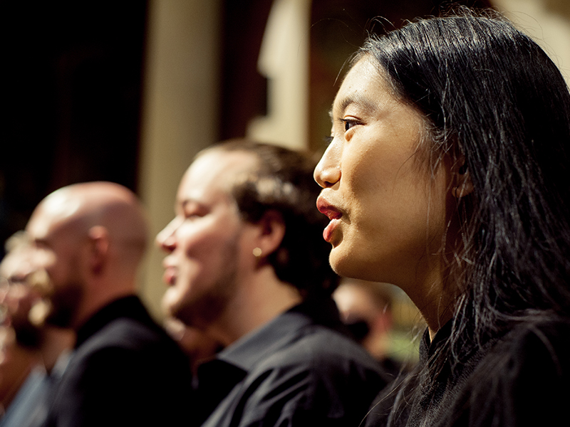 This is a close-up view of three students in concert attire singing onstage with the rest of the University Chorus. 
