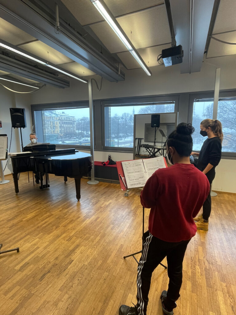 Two students are seen rehearsing with an instructor at the piano. 