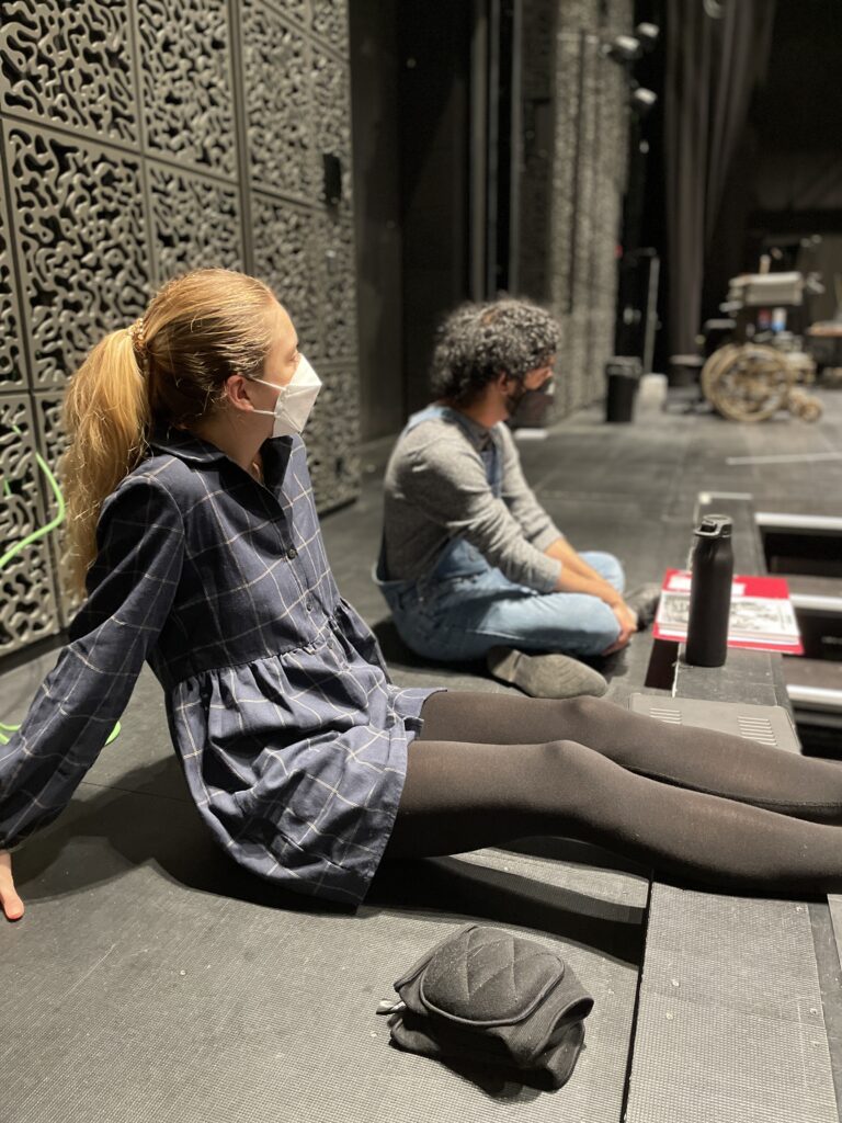Third of three pictures shows two students sitting on the floor looking at a rehearsal taking place onstage indoors