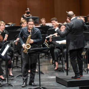 Student saxophonist performs onstage indoors with an orchestra led by a conductor in the background.