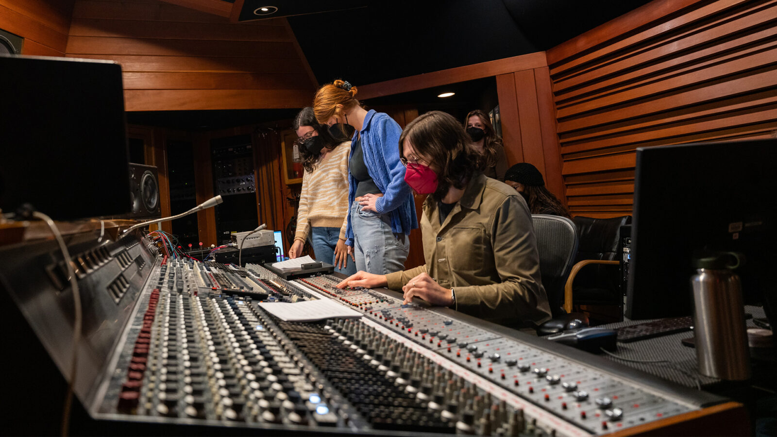 Photo of popular music students working around a professional mixing board at a sound studio. 