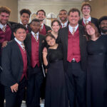 Photograph of college-aged choral singers in black concert attire smiling together inside a cathedral.