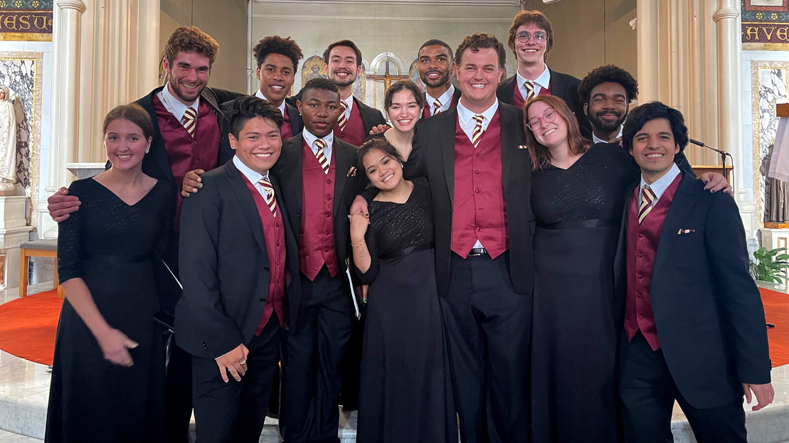 Photograph of college-aged choral singers in black concert attire smiling together inside a cathedral. 