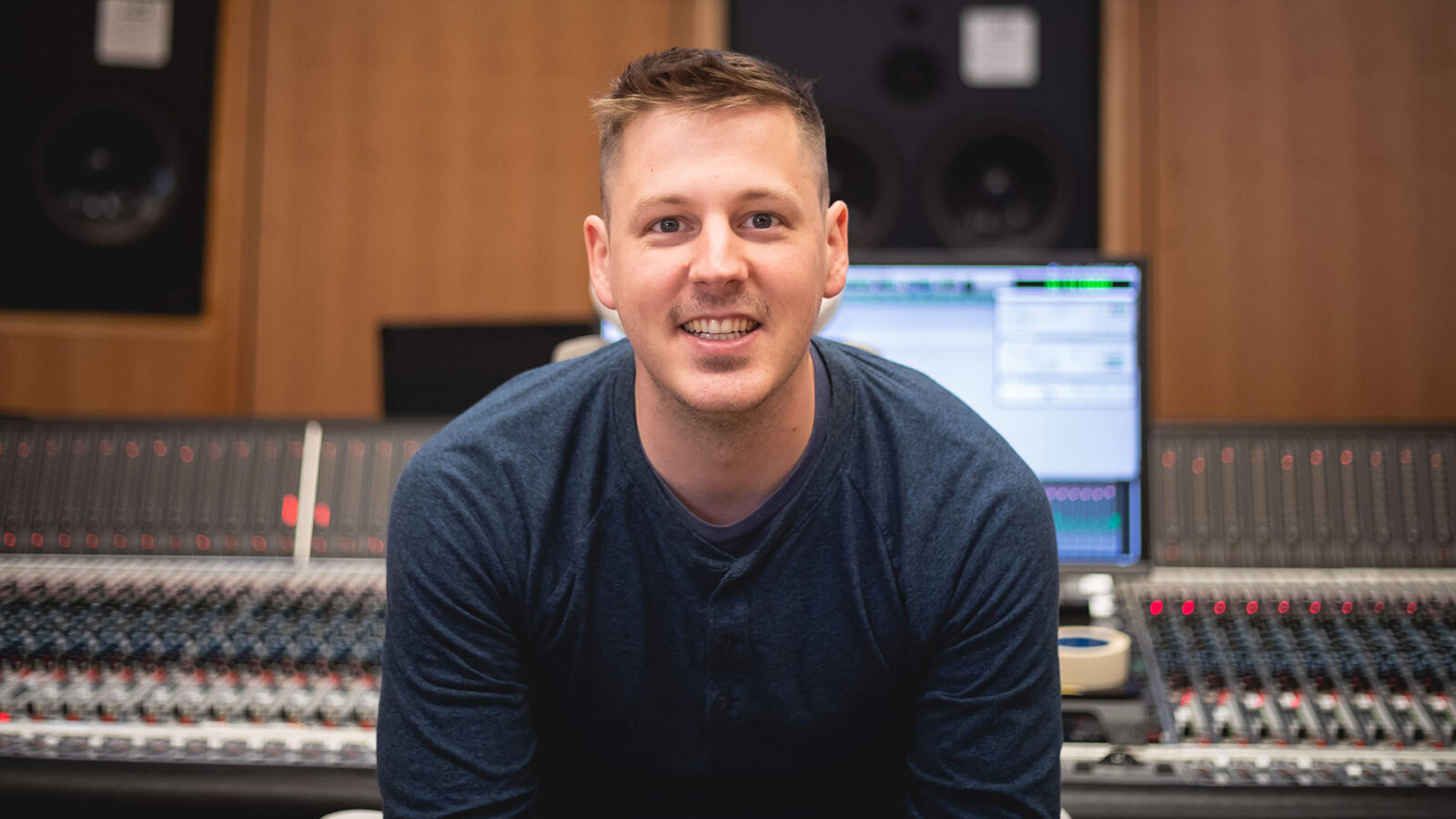 Photo of Christian Amonson smiling in front of a mixing console in a recording studio. 