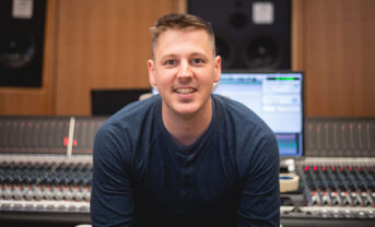 Photo of Christian Amonson smiling in front of a mixing console in a recording studio.
