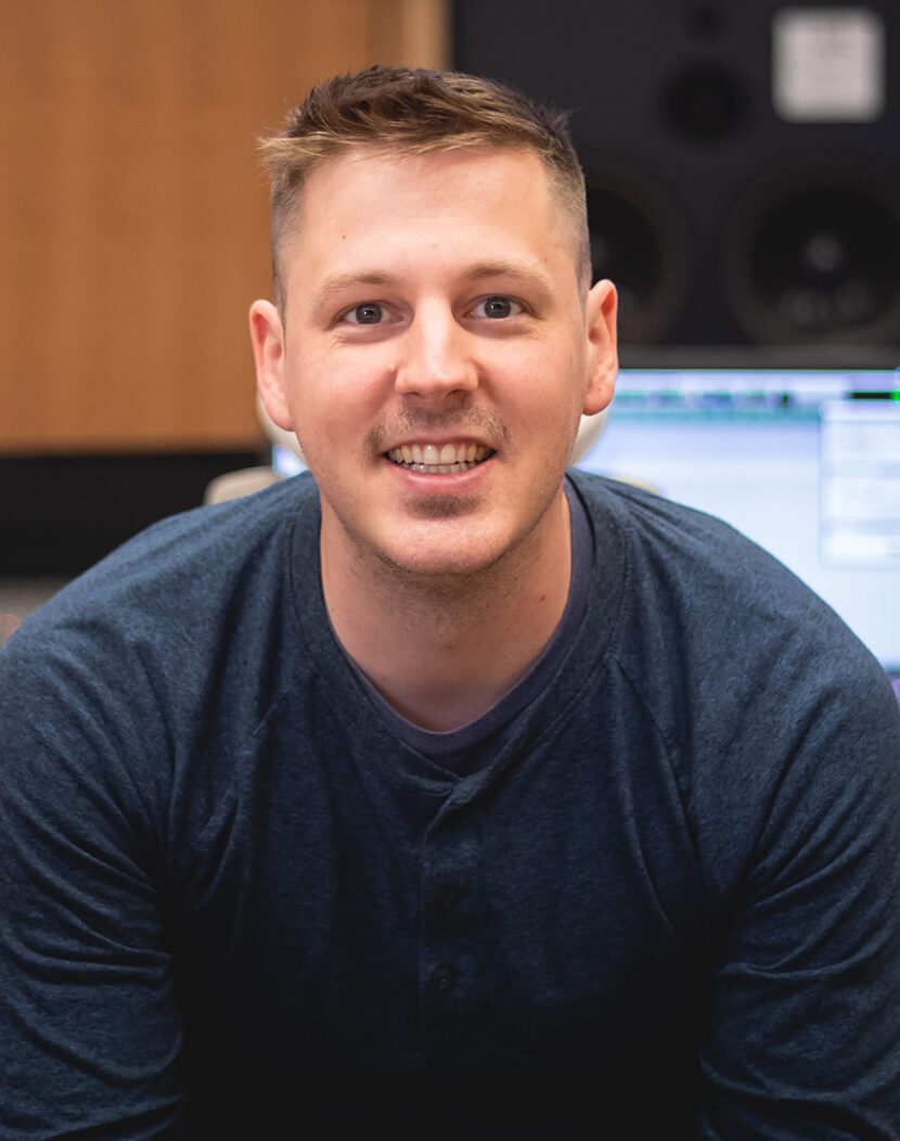 Photo of Christian Amonson smiling in front of a mixing console in a recording studio.