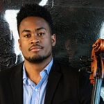 Photo of Ismael Ariel Guerrero Bombut holding his cello in front of a downtown city wall.