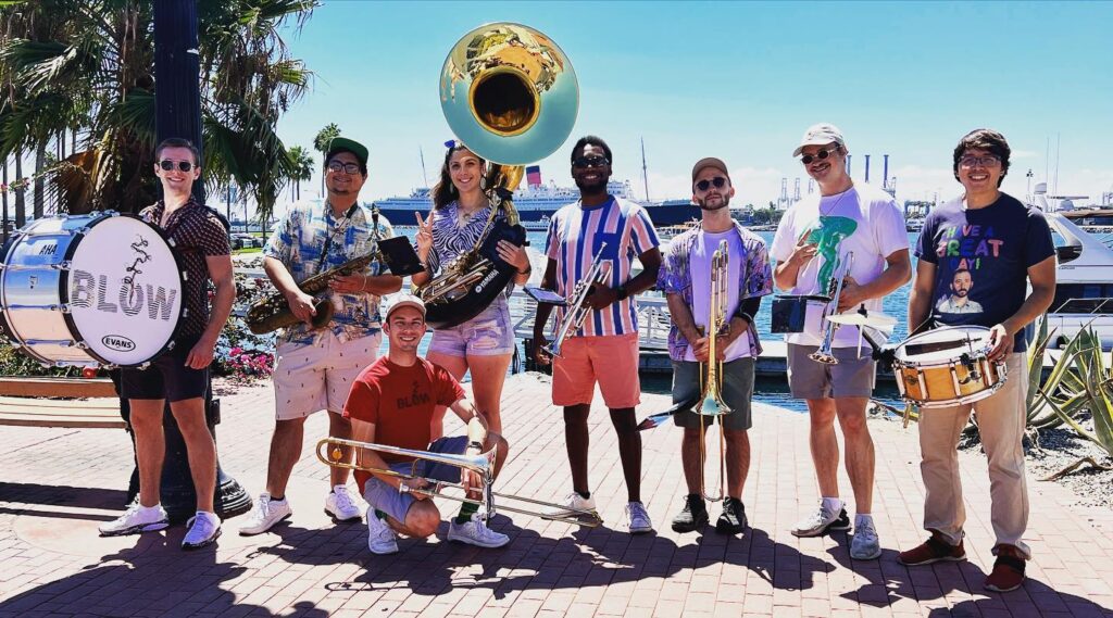 Photo of a brass band holding their instruments outdoors by a pier in the afternoon sun.