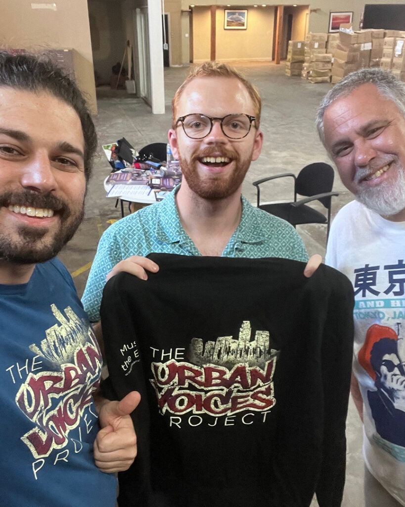 Photo of smiling people holding up black shirts that read: "The Urban Voices Project".