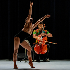 Photo of a cellist performing on stage with a dancer in a black unitard in front of them.