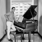 Black and white photo of composer Juan Pablo Contreras standing in front of a piano indoors.