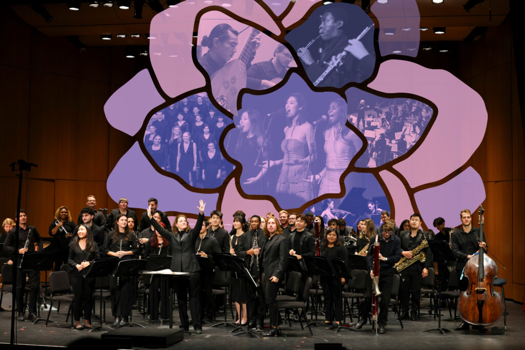 A symphony orchestra bows after a performance on a concert stage, in front of an illustration of a purple flower.