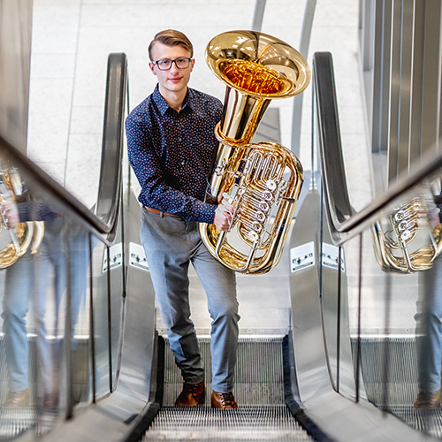 Student wearing glasses and carrying a tuba ascends an escalator inside an office building.