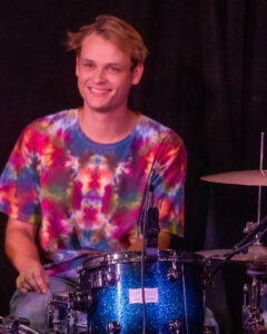 A drummer wearing a tie-dye shirt smiles behind a drum kit.