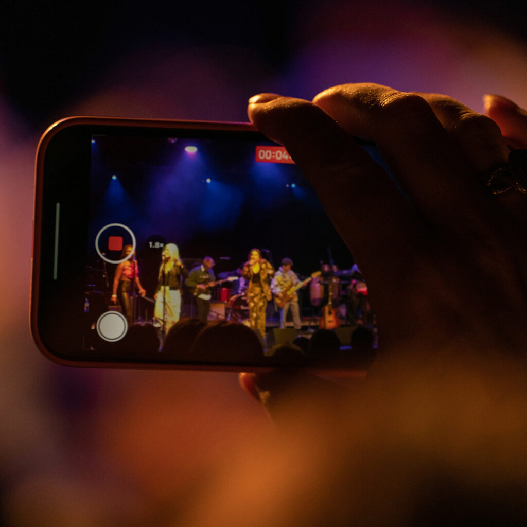 A hand holding a smartphone video records a popular music concert.