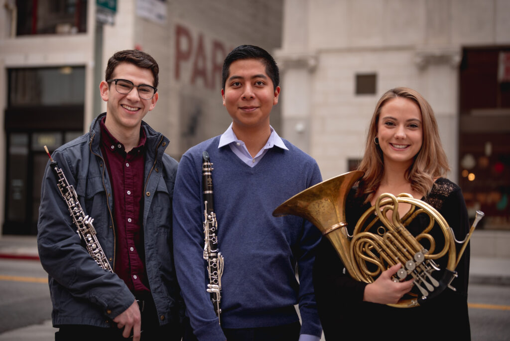 Three students holding musical instruments and smiling outdoors in downtown Los Angeles.