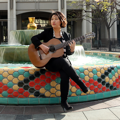 Photo of a classical guitarist playing her instrument outside in a city environment.
