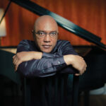 Photo of composer Billy Childs sitting in front of a piano.