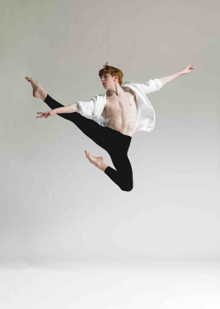 Photo of a dancer wearing black leggings and a white shirt leaping into the air.