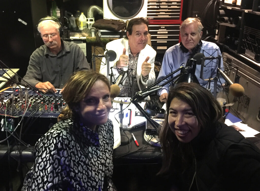 Photo of radio hosts with recording equipment smiling backstage at a classical music concert.