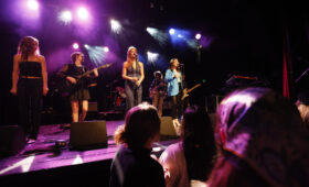 Students singing and playing onstage under colored lights for a crowd.