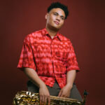 Saxophonist with dark hair wearing a red shirt and holding his instrument in his lap.