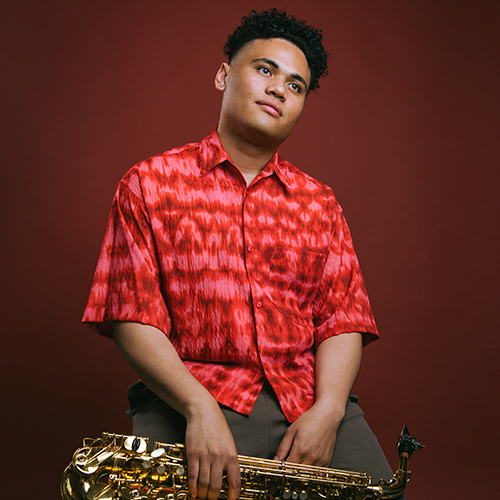 Saxophonist with dark hair wearing a red shirt and holding his instrument in his lap.