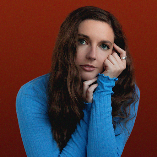 Singer Katie Stump in a blue sweater poses in front of a red wall.