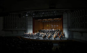 Student symphony performs onstage, led by a female conductor.