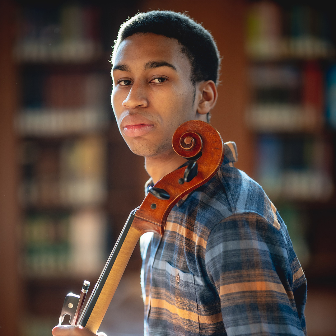 Cellist Quenton Blache holding his instrument and sitting in a library.
