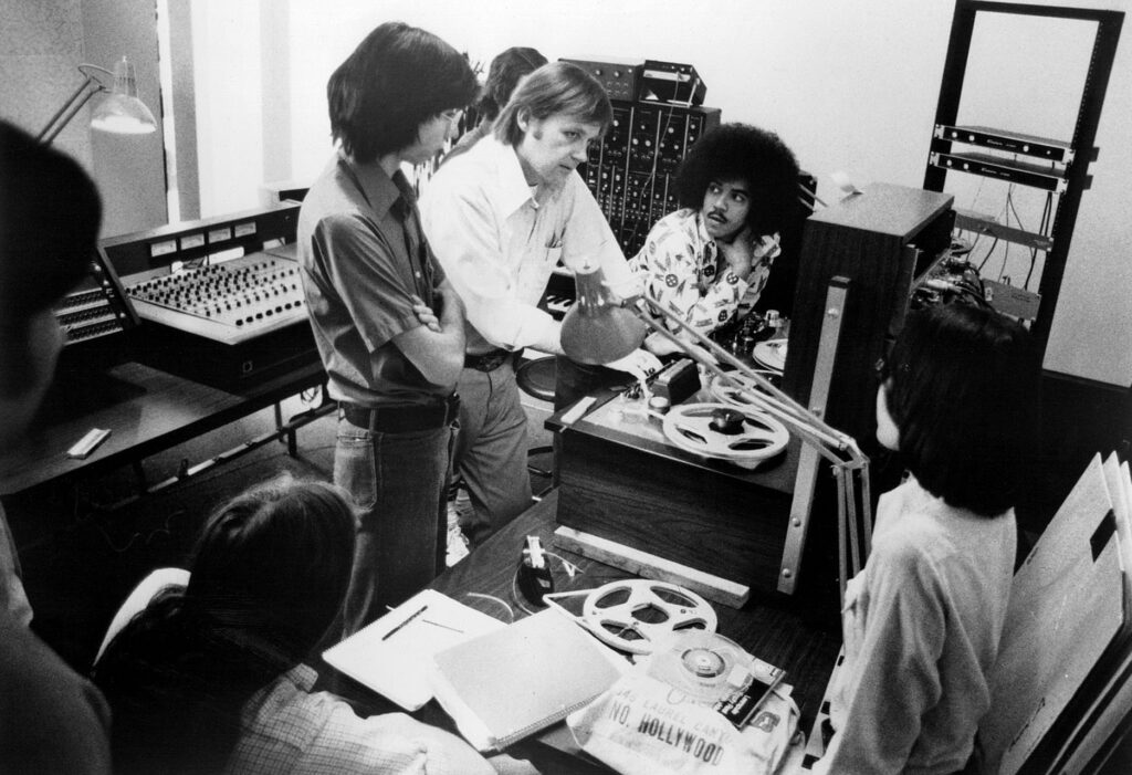 Black and white photograph of a music instructor surrounded by electronic equipment from the 1970s, instructing a classroom of students.