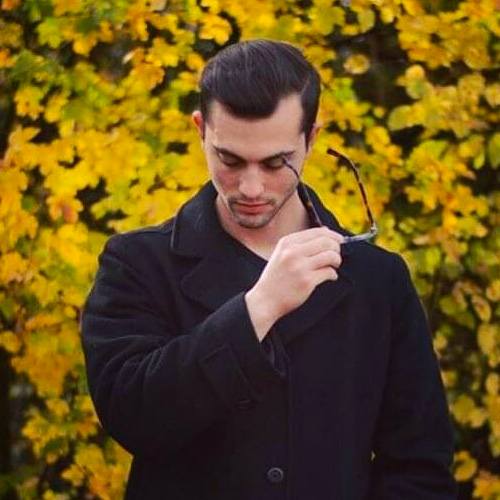 USC Thornton alumnus Daniel Newman-Lessler standing in front of yellow bushes wearing a black coat and taking off glasses.