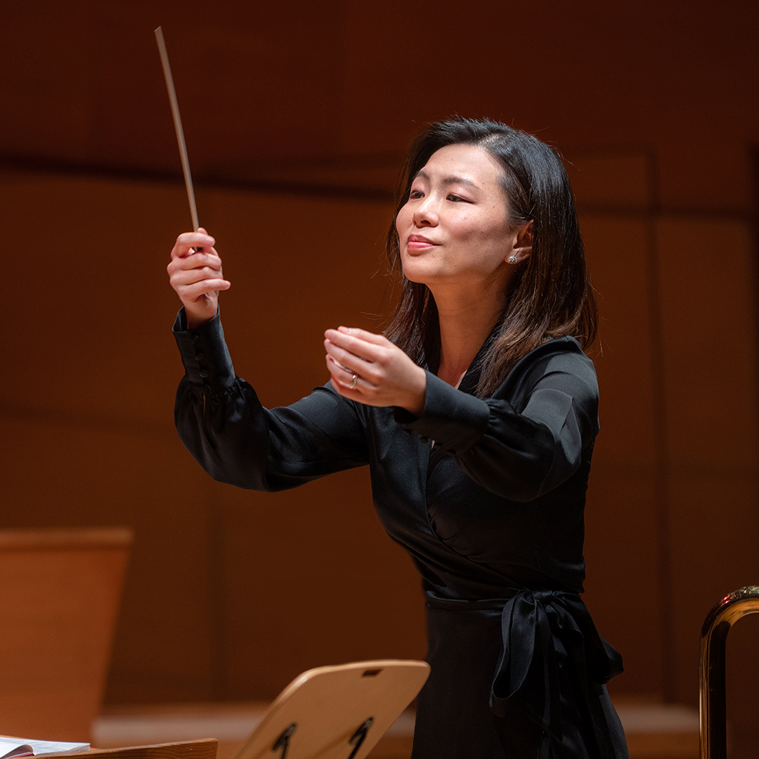 Jenny Wong in concert black attire conducting a choir from a podium with a baton.