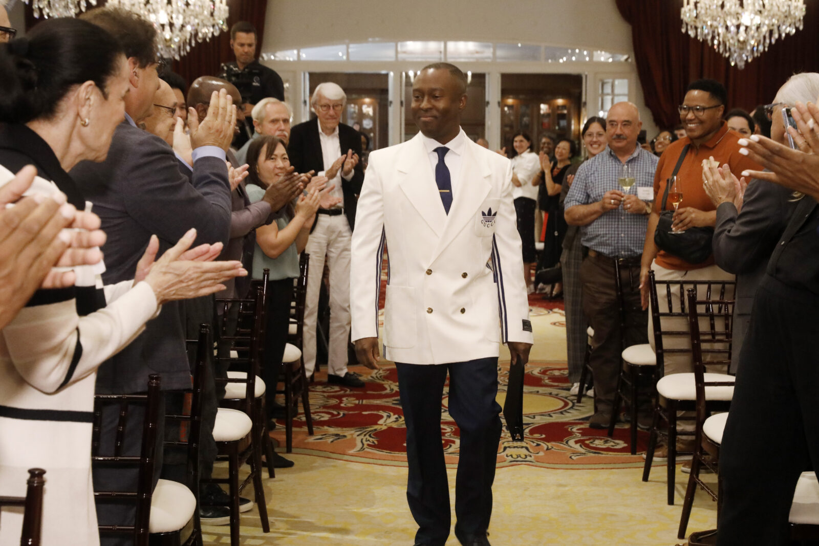 Dean Jason King dressed in a white blazer walks down the aisle during a ceremony in a ballroom. 