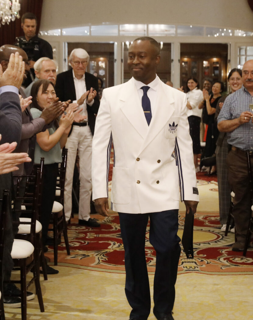 Dean Jason King dressed in a white blazer walks down the aisle during a ceremony in a ballroom.