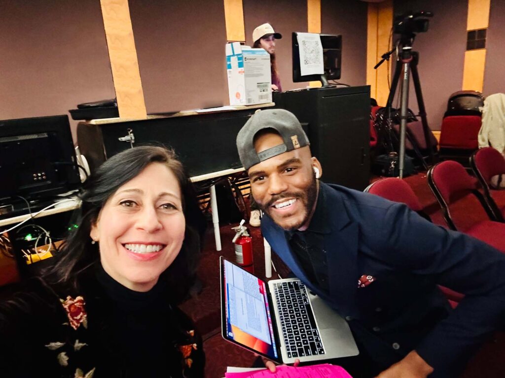 Two smiling people in a room looking into the camera with one holding a laptop.