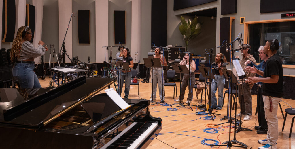 A choral ensemble recording a popular music song led by a conductor on a podium in a large recording studio surrounded by microphones.